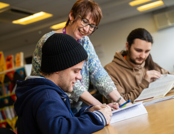 A smiling teacher standing over two students at a table, referencing an open textbook