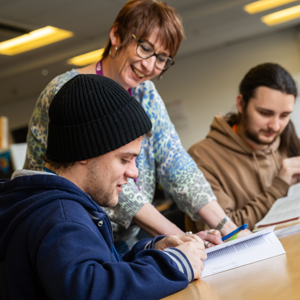 A smiling teacher standing over two students at a table, referencing an open textbook