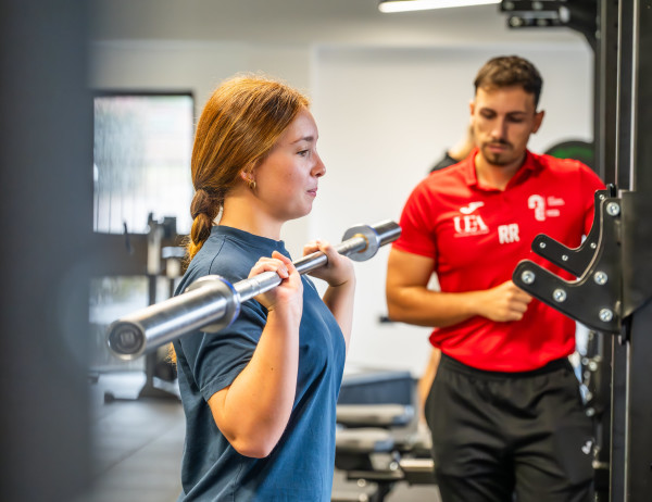 A student lifting a weight, with a coach standing behind. 