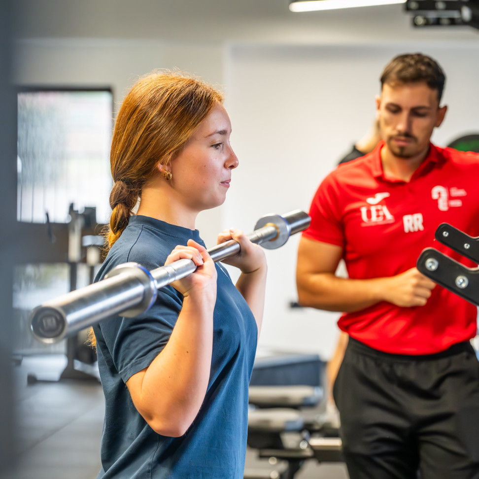A student lifting a weight, with a coach standing behind. 