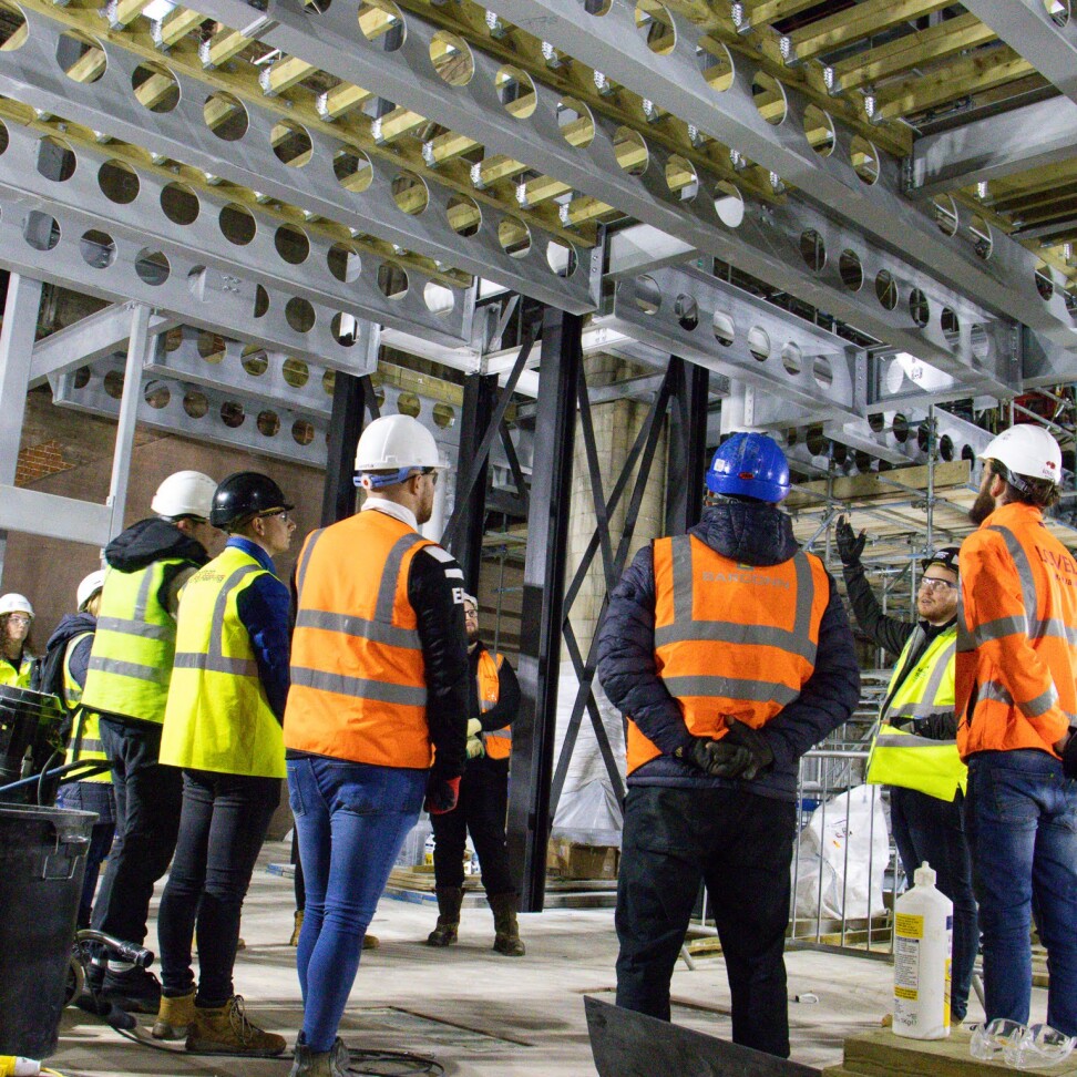 A group of students are at a construction site, standing around a lecturer.