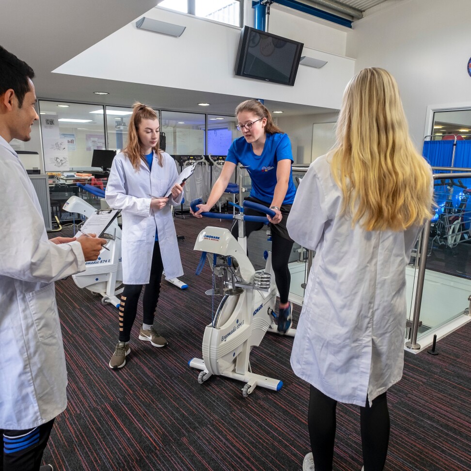 Three Sport, Health and Exercise students stand in lab coats, holding clipboards. They are assessing another student, who rides an exercise bike,