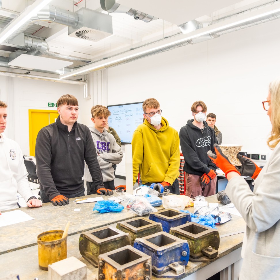 A Civil Engineering classroom. Students are gathered around a lecturer, who is holding concrete.