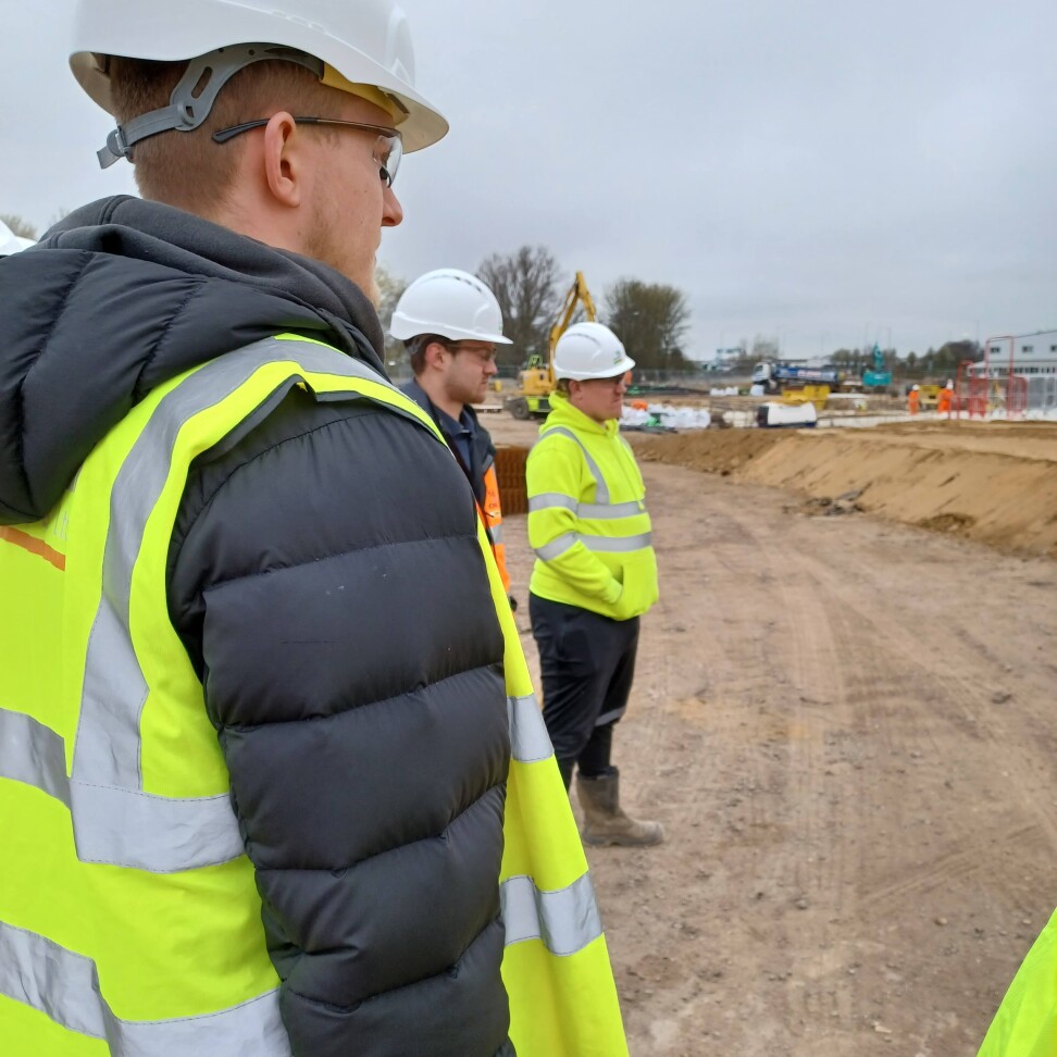 Three students are standing at a construction site, wearing hi-vis jackets and hard-hats. They are looking away from the camera.