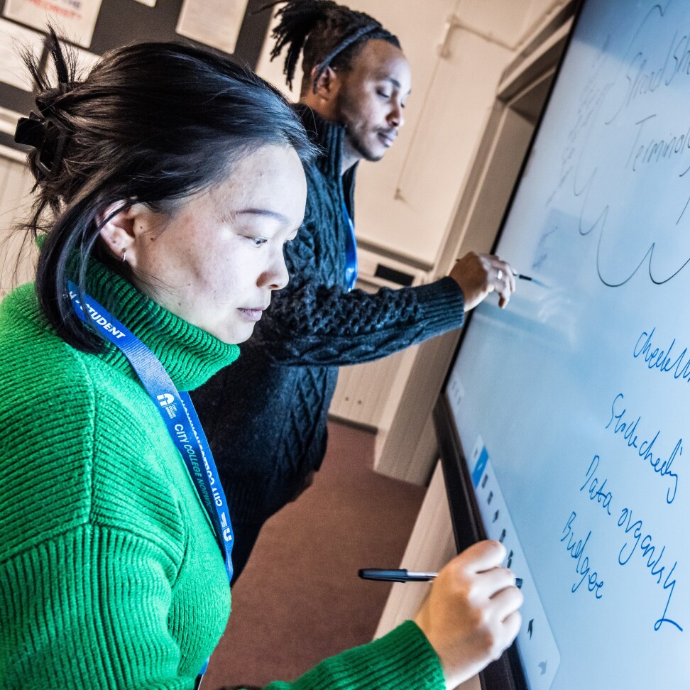 Two Digital Technologies students standing at the front of a classroom, writing on an interactive whiteboard.