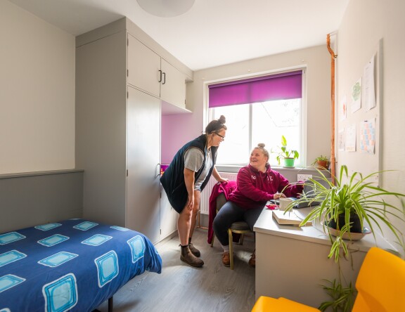 Two students in Easton accommodation. One student is sat at a desk in their room, and the other is standing and chatting to them.