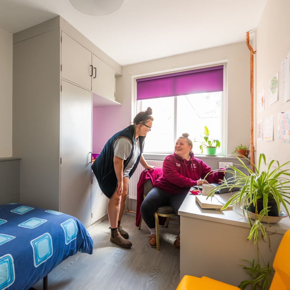 Two students in Easton accommodation. One student is sat at a desk in their room, and the other is standing and chatting to them.