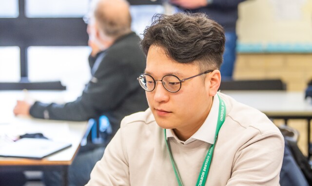 A Higher Education student sitting on their laptop at a table. A teacher stands in the background.