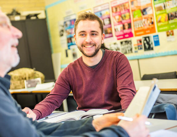 A Higher Education student sat at a table, smiling. Another student is sat close by, book in hand.