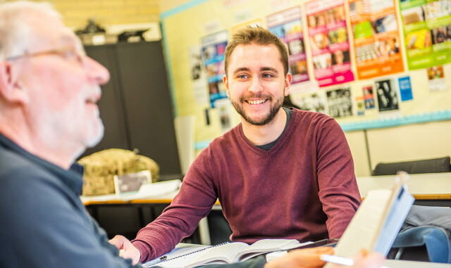 A Higher Education student sat at a table, smiling. Another student is sat close by, book in hand.