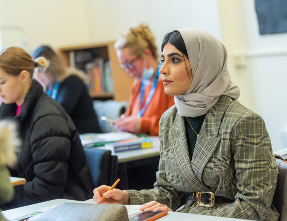 A student sat at a desk, pencil in hand, looking towards the front of the classroom. Other students work in the background.