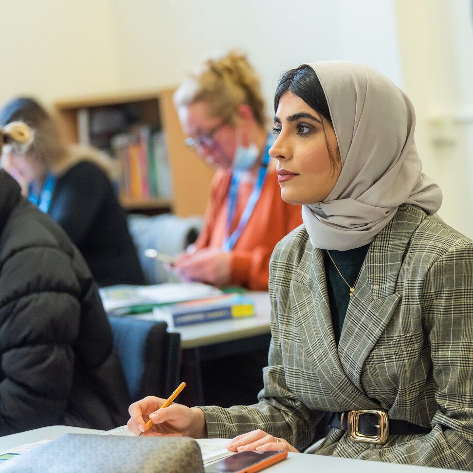 A student sat at a desk, pencil in hand, looking towards the front of the classroom. Other students work in the background.