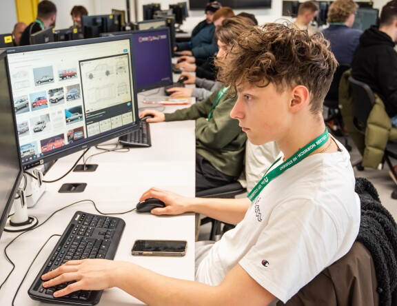 A computer room. A Higher Education student is typing at a computer; other students are working at computers in the background.