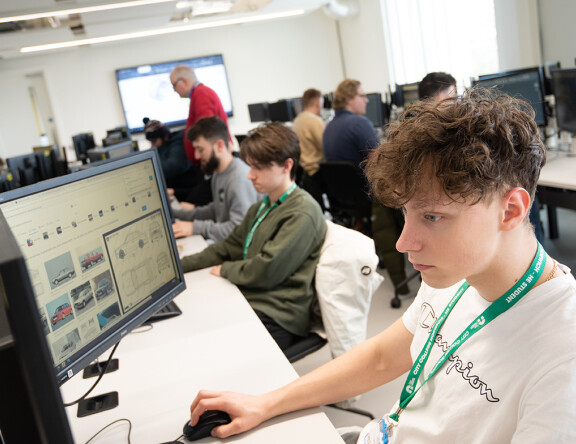 A Higher Education student in a computer room, working at a computer. A teacher can be seen standing in the background, while other students also work at computers.