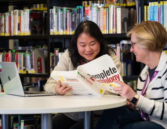 An Access to Higher Education: Humanities and Social Sciences student is sat at a table in the library. They are discussing a book with a teacher, who sits next to them.