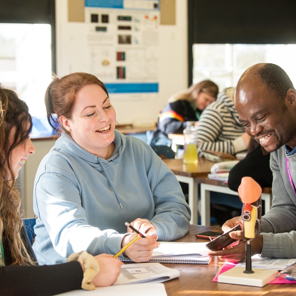 Two Access to Higher Education: Science students are sat at a table, with a teacher. A teacher is showing an anatomical model to the students.