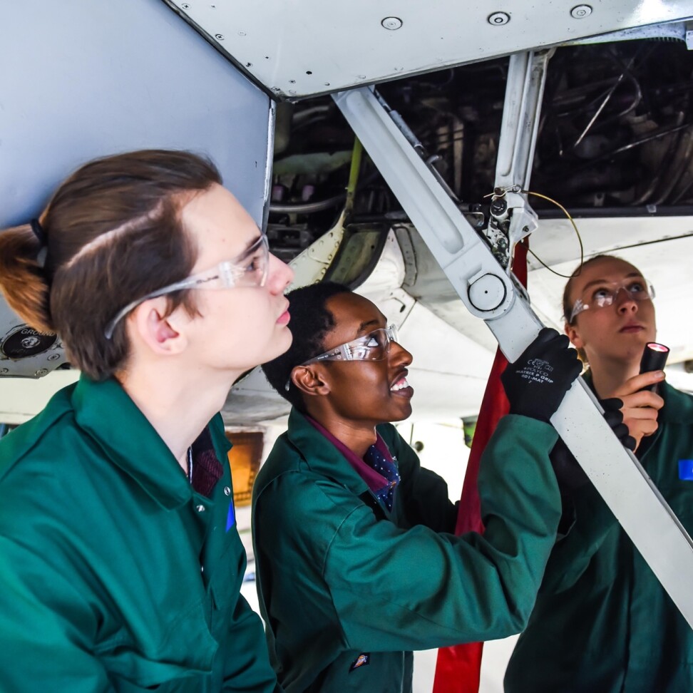 Three Aviation students are gathered under the body of a Boeing 737 plane, staring up into the interior. One is shining a torch.