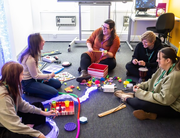 A group of BA Hons Additional Needs and Disabilities: Children and Young People students are sat in a circle on the floor. A teacher sits in the middle, and is demonstrating the use of sensory toys.  