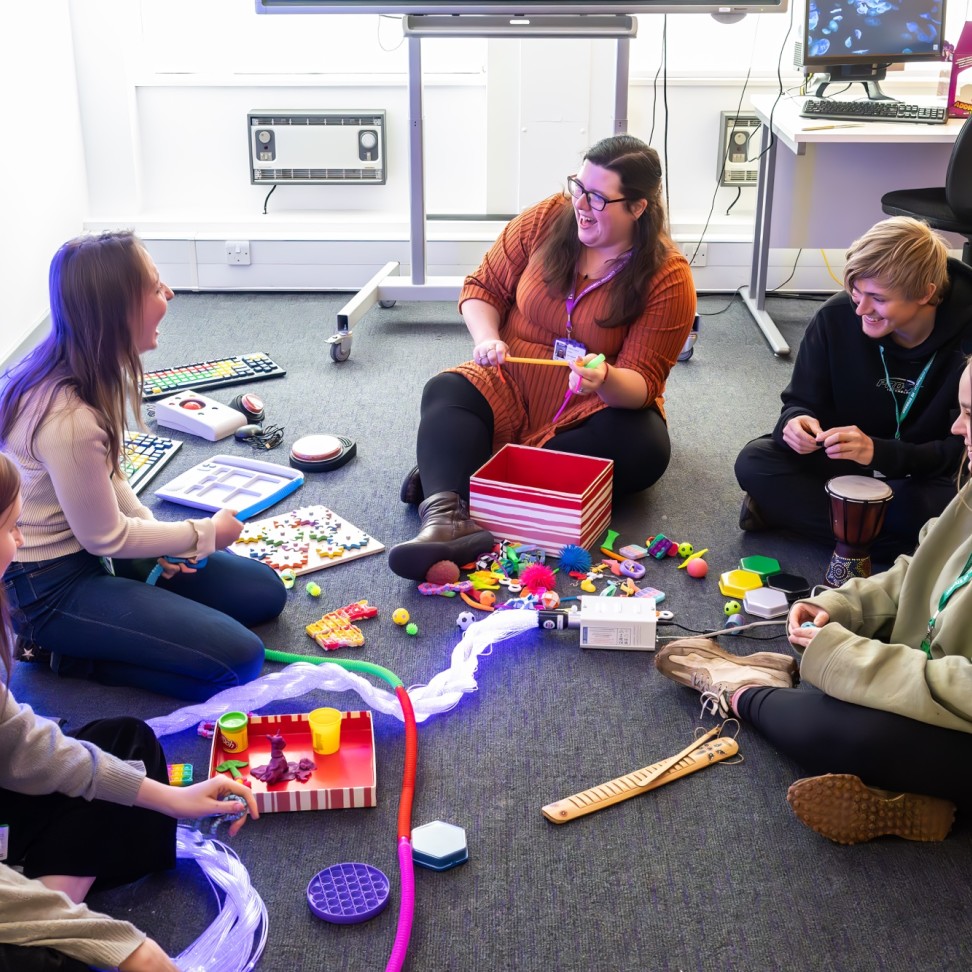 A group of BA Hons Additional Needs and Disabilities: Children and Young People students are sat in a circle on the floor. A teacher sits in the middle, and is demonstrating the use of sensory toys.  