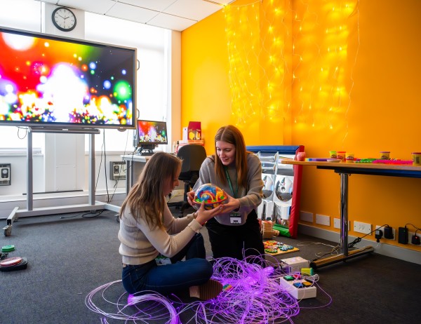 Two BA Hons Additional Needs and Disabilities: Children and Young People students are sat on the floor of a classroom. They are holding a sensory toy.