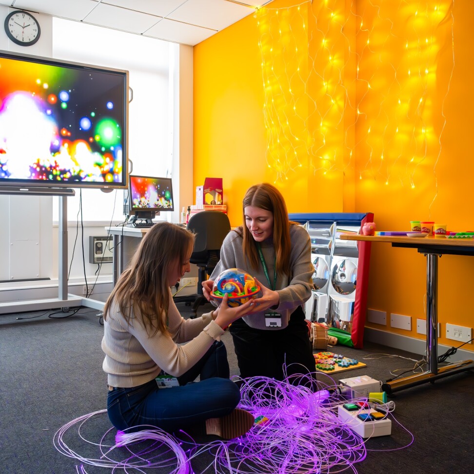 Two BA Hons Additional Needs and Disabilities: Children and Young People students are sat on the floor of a classroom. They are holding a sensory toy.