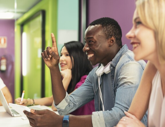 Three Leadership Management students are sat at a desk, smiling. One has their hand up.