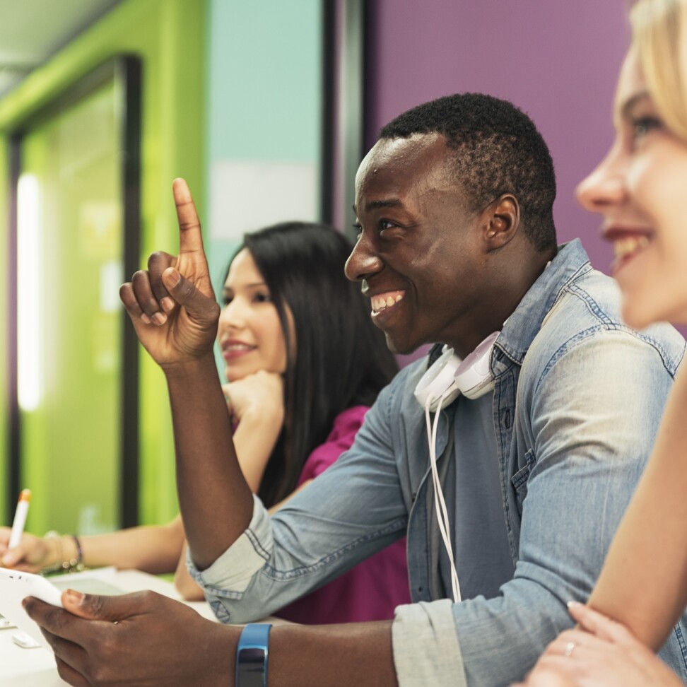 Three Leadership Management students are sat at a desk, smiling. One has their hand up.