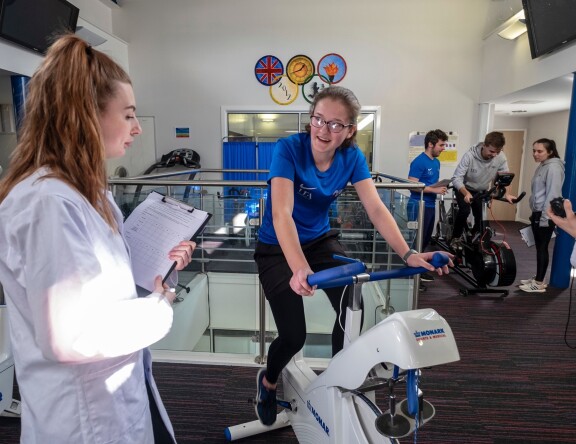 Some Sport, Health and Exercise Science students are working on exercise bikes. One student observes with a clipboard, as another student is on the bike.