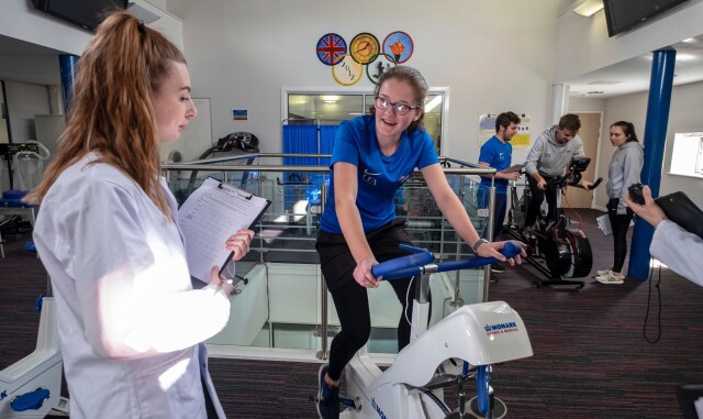 Some Sport, Health and Exercise Science students are working on exercise bikes. One student observes with a clipboard, as another student is on the bike.