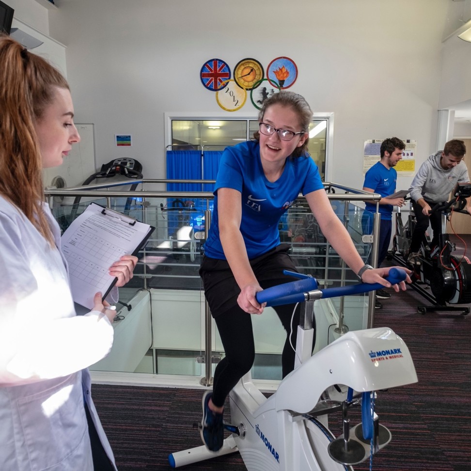 Some Sport, Health and Exercise Science students are working on exercise bikes. One student observes with a clipboard, as another student is on the bike.