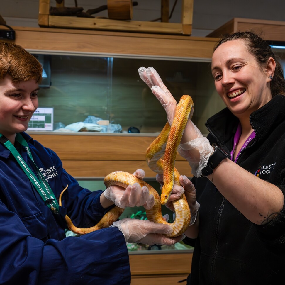 A Zoology student is holding a snake, with the assistance of a teacher. Both are smiling. 