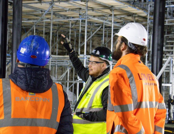 Construction Management students are at a construction site. They are watching a teacher, who is gesturing towards the scaffolding.