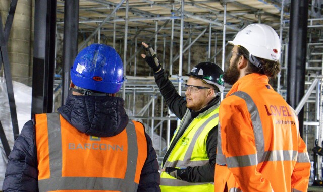 Construction Management students are at a construction site. They are watching a teacher, who is gesturing towards the scaffolding.