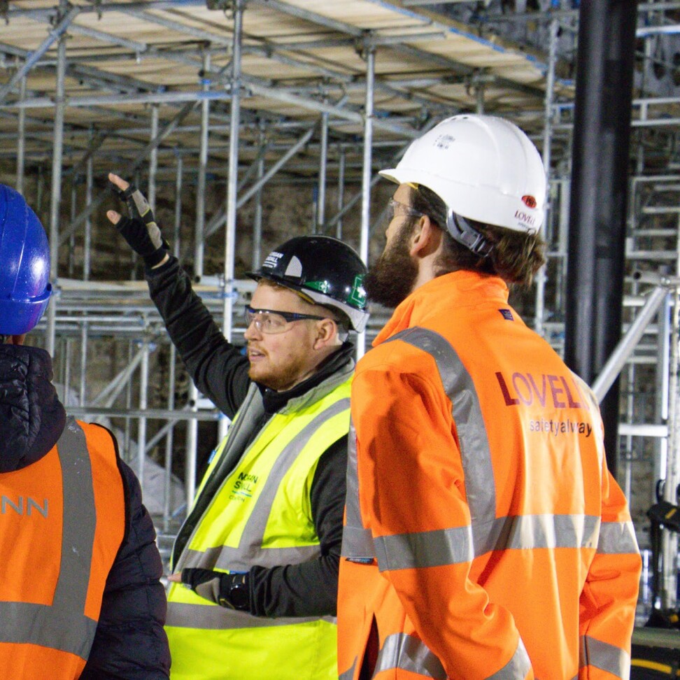 Construction Management students are at a construction site. They are watching a teacher, who is gesturing towards the scaffolding.