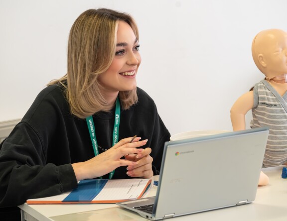 A Childhood Studies student is sat at a table, with their laptop. Sitting at the table is a model baby. The student is smiling and looking towards the front of the classroom.