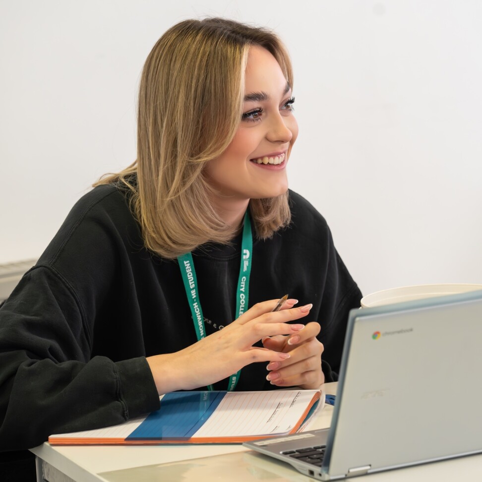 A Childhood Studies student is sat at a table, with their laptop. Sitting at the table is a model baby. The student is smiling and looking towards the front of the classroom.