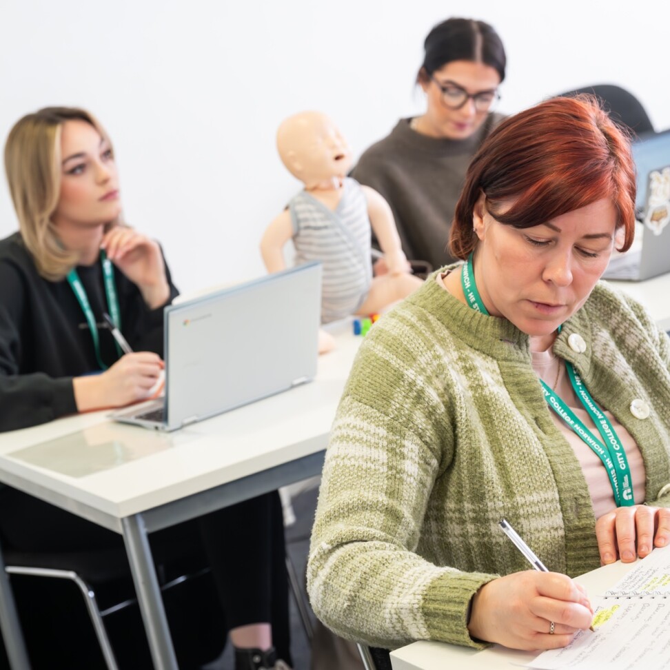 Three Childhood Studies students are sat at desks, typing on laptops or writing in notebooks. A model baby is sat on the desk.