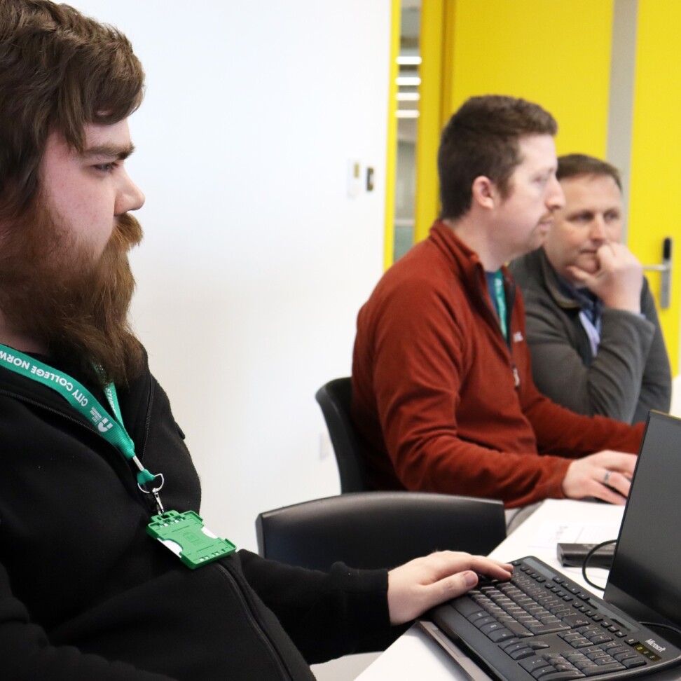 Two HNC Electrical and Electronic Engineering for England students are sat at computers. A teacher is assisting one of the students, sitting beside them.