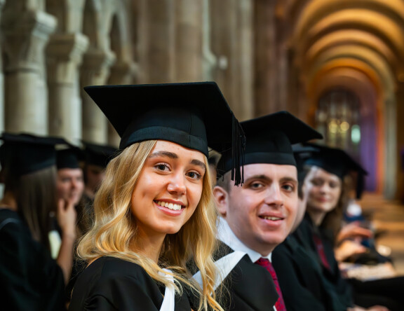 Students are sat in rows at the graduation ceremony. Two students look at the camera, smiling.
