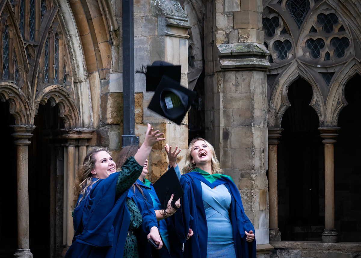 Three Higher Education students at graduation. They are stood outside the Cathedral, and are throwing their caps in the air.