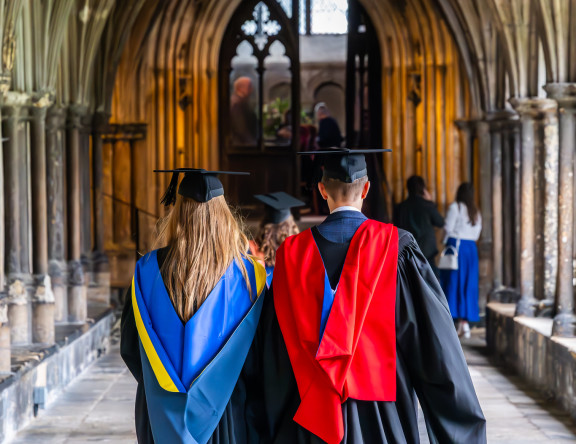 Two graduates are walking away from the camera, along a corridor in the Cathedral.