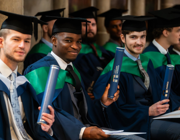 A group of Higher Education graduates, sat in rows inside the Cathedral. They are smiling and holding up their certificates.