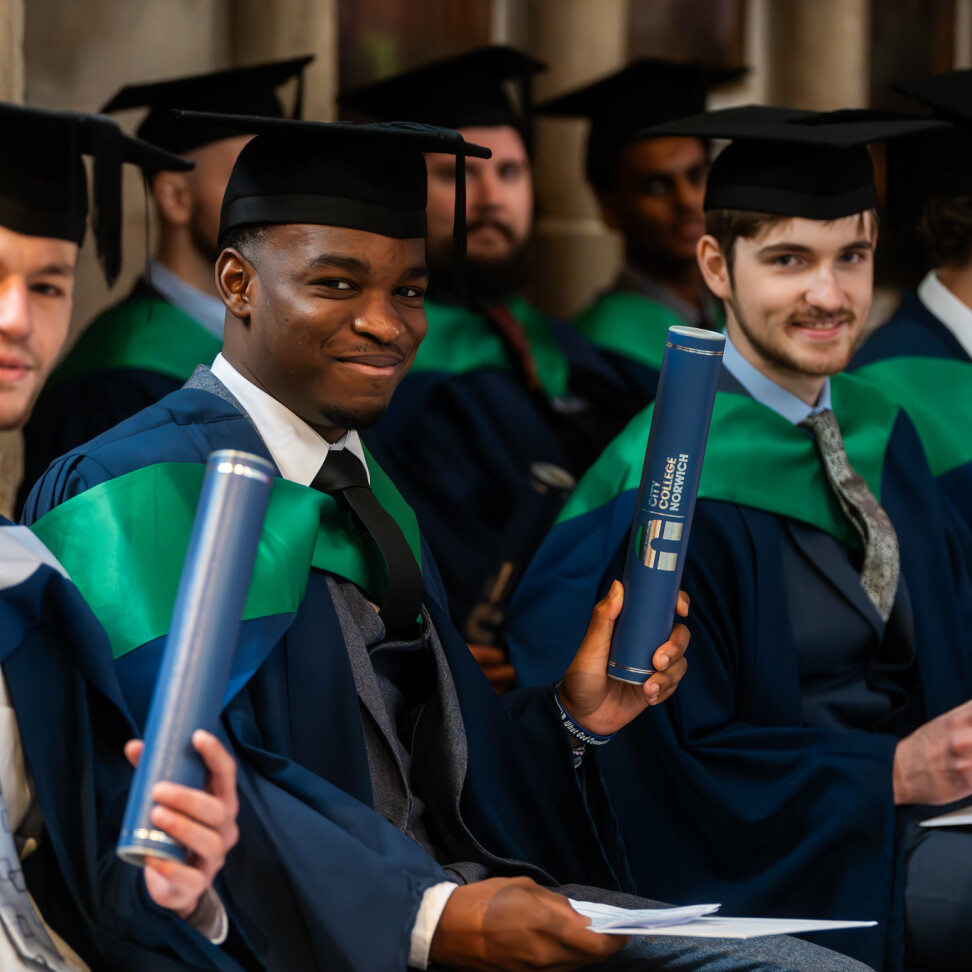 A group of Higher Education graduates, sat in rows inside the Cathedral. They are smiling and holding up their certificates.