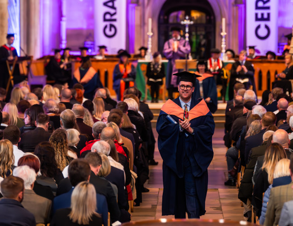 This image depicts the graduation ceremony and stage, showing the audience from the back. A student is walking up the middle aisle, certificate in hand.