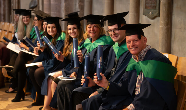 A group of Higher Education graduates sat in a row inside the Cathedral. They are smiling and holding their certificates.