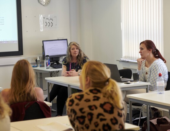 Taken from the back of a classroom. The teacher sits at a desk, leading a group discussion. Students can be seen looking at the board.