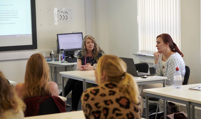 Taken from the back of a classroom. The teacher sits at a desk, leading a group discussion. Students can be seen looking at the board.