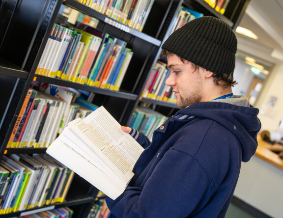 A student is in the library. They are standing in front of a bookshelf, reading a book.