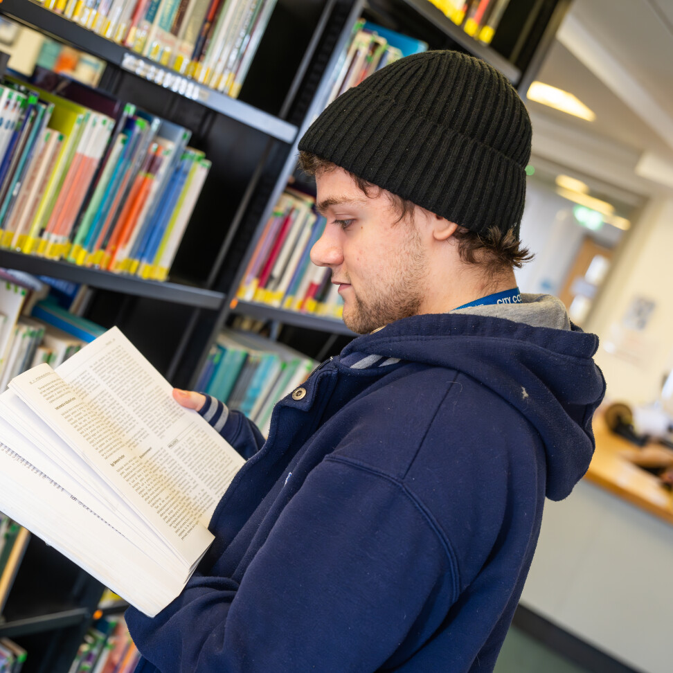 A student is in the library. They are standing in front of a bookshelf, reading a book.