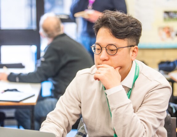 A Higher Education student is sat at a table, working on a laptop. A teacher can be seen standing in the background.